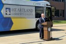 A man stands speaking at a podium, with a large bus behind him. The Connect Transit bus has a hawk on it and says Heartland Community College on the windows.