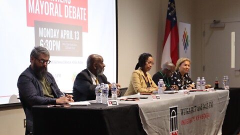 Candidates sit at a table during an Athens mayoral debate, with an event slide projected on a screen behind them.