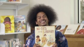 Mychal Threets, the host of "Reading Rainbow," is seen smiling and holding a book titled, "No Cats in the Library."