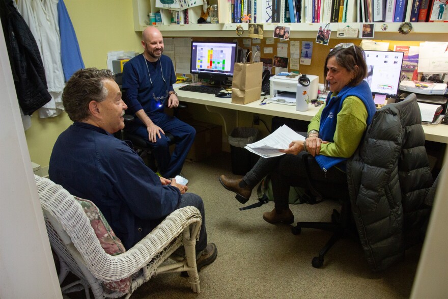 A photo of three people, two men and a woman, sitting in a small office and smiling while talking. On the wall behind them are books, photos and computer screens.