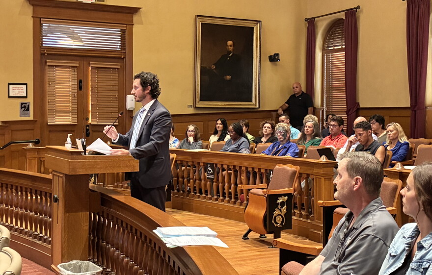 A man in a suit speaks at a podium in a courtroom filled with seated people. A large portrait hangs on the wall behind him. Papers are on the podium and benches.