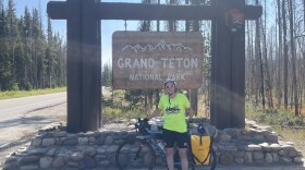 Friederike Benning stands smiling with her bike in front of the Grand Teton National Park sign