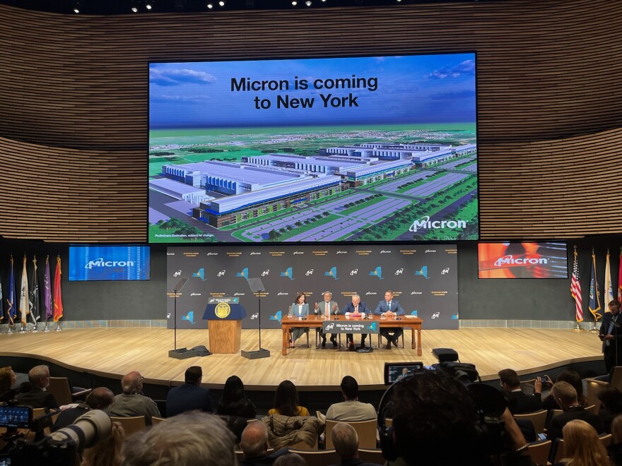 Gov. Cathy Hochul, Sen. Chuck Schumer, Micron CEO Sanjay Rehrotra and Onondaga County Executive Ryan McMahon sit on a stage underneath a large screen reading "Micron is coming to New York."