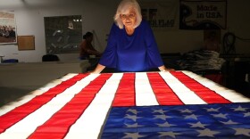 An elderly woman with white hair and glasses looks at an American flag illuminated from underneath by a light table.
