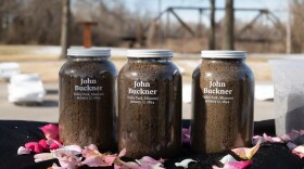 Three jars, each filled with dirt, sit on a dark tablecloth surrounded by pink and white flower petals during a soil-collection ceremony for 1894 lynching victim John Buckner. White letters on the jar write "John Buckner, Valley Park, Missouri January 17, 1894." The soil collected at the 2022 ceremony became part of a memorial exhibits in several states, including the Griot Museum. 