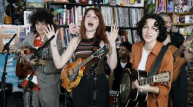 Three musicians holding guitars perform behind an office desk with two more musicians in the background smiling.