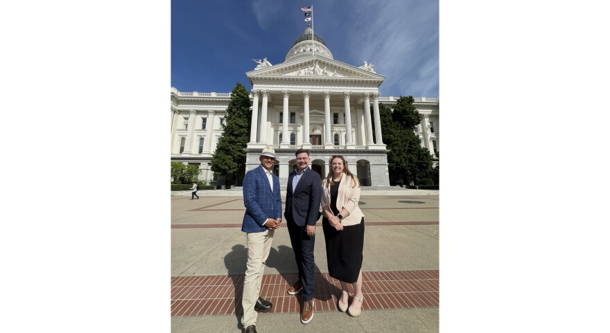 Three people stand in front of the California State Capitol on a sunny day. 
