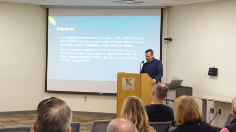 Multiple heads of attendees sit at the bottom of the photo, which takes place in a white walled room. A speaker stands at a light brown podium in front of the audience, he looks down as he speaks into the mic in front of him. Next to him, further to the right of the image, is a large projector screen with his presentation. The presentation goes over what an Impasse in negotiations means, with several bullet points going how they got there and what it means for the bargaining process.  