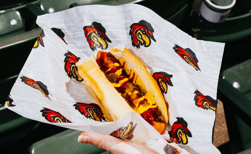 An arm holding a hot dog covered in ketchup and mustard in a bun on a paper with the Red Wings logo