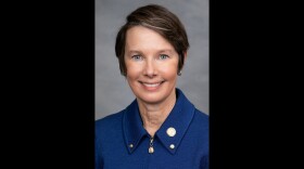 A close-up photo of a woman with short hair and a blue-collared top. Black bars on side of image. 