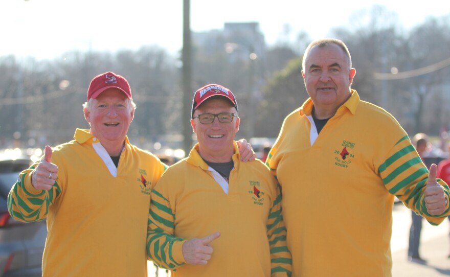 Three smiling men wearing yellow rugby shirts with green-striped sleeves and red caps stand together outdoors, giving thumbs up in a sunny setting. Trees and cars are visible in the blurred background.