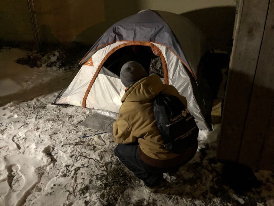 A person crouches outside a tent where people are living. There is snow on the ground.