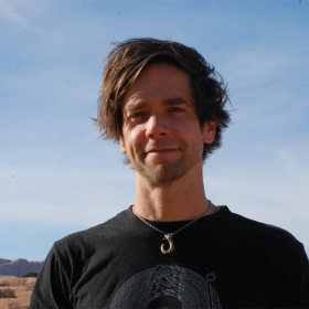A man with medium-length brown hair stands outside. He wears a black shirt and a hook necklace. He looks at the camera and smiles. 