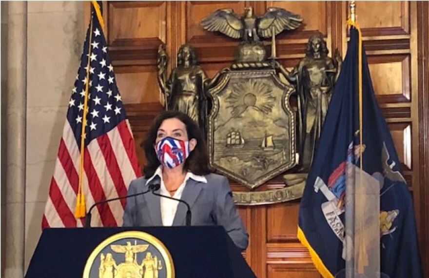 Gov. Kathy Hochul, wearing a gray suit, white shirt and Buffalo Bills face mask, at a podium