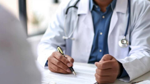 A doctor, with a stethoscope around his neck, writes on a paper on a desk