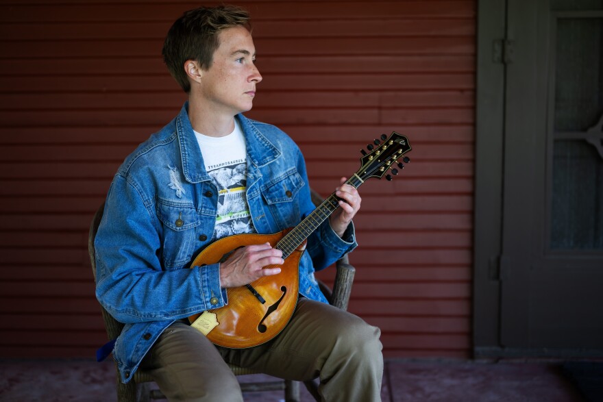 a man sitting on a chair on a porch, playing a new-looking mandolin and gazing off camera