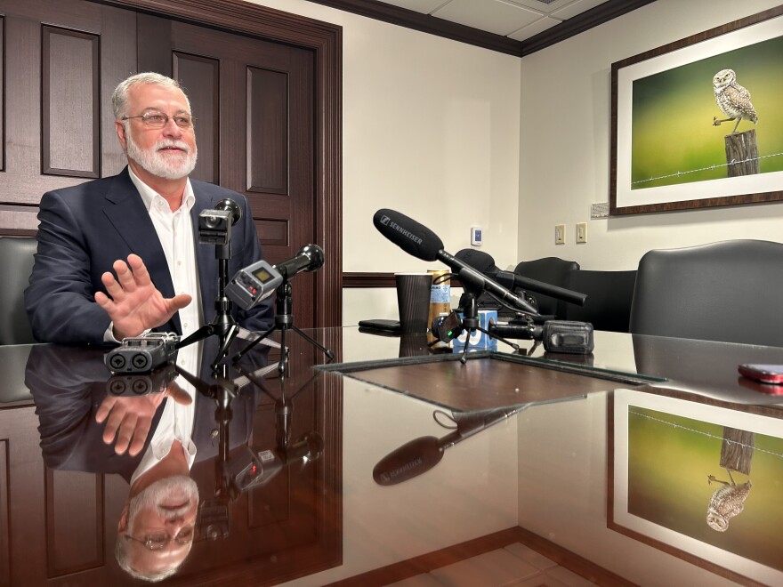 Man with gray hair and beard wearing a dark suit jacket and sitting at a table to the left with microphones in front of him