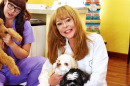 Two women sit on a hard-wood floor. One woman wears a white lab coat and has long blonde hair, she smiles at the camera while holding two dogs. The other woman wears purple scrubs, has long black hair and glasses, and holds a dog. 