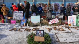 people stand on a street with signs strewn about; a picture of a man l ies in a wreath. "alex Peretti" is spelled out in rocks