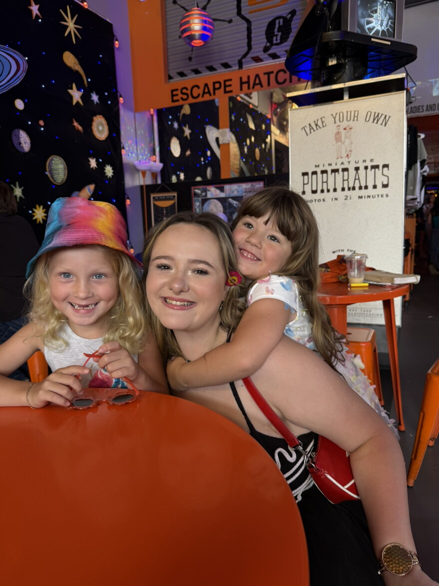 A young woman smiling alongside two young children in a colorful, space-themed room.