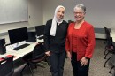 Two women standing in a classroom between rows of students desks and computers. The women on the left is wearing a dark suit with white hijab, the woman on the left is wearing a red top and jacket with blank pants.