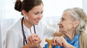 A nurse wearing white scrubs and a stethoscope holds the hand of an elderly woman, wearing a blue blouse and beige vest.