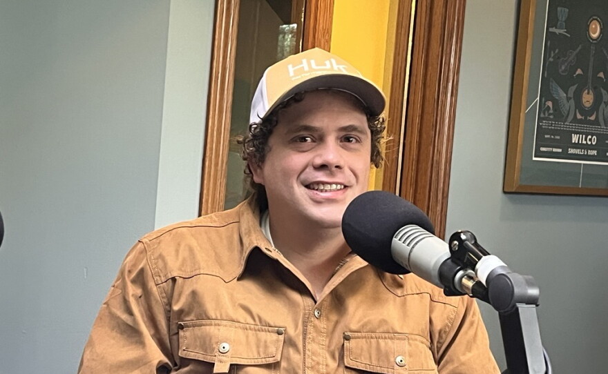 A man in a tan jacket and yellow baseball cap sits behind a microphone in the KAXE studio.