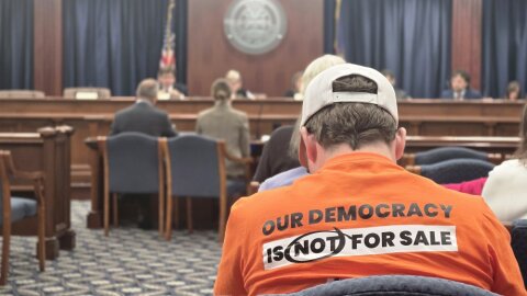 A person in an orange shirt reading "our democracy is not for sale" sits facing away from the camera as a meeting proceeds at the front of a carpeted room.