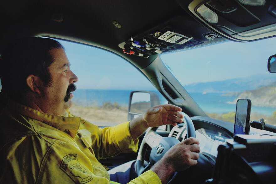 A man in a yellow firefighting jacket drives on a highway with the ocean behind him. 