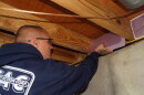 An over-the-shoulder image of a bald man in a navy blue hoodie placing a piece of purple foam insulation in between the beams of a ceiling. 