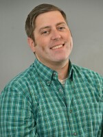 A man wearing a green shirt sits smiling for the camera on a gray background.