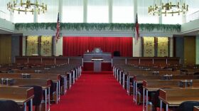 The inside of the NC House chamber showing wooden desks, red carpet and a podium in the background in front of the speaker's dais. A US flag is hanging on the left and the NC state flag on the right.