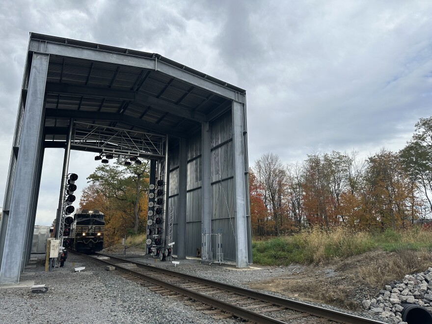 A Norfolk Southern train rushes towards a large metal shed-like structure with lights inside.