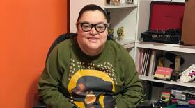 A smiling woman with her dark hair pulled into a ponytail. She is wearing glasses, a green sweatshirt with an image of a woman, white pants, and green shoes. She is using a wheelchair. 