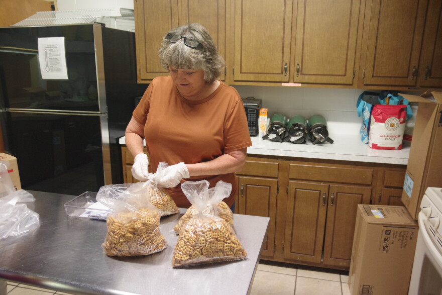 A woman with medium length hair in a short sleeve dark orange t-shirt twists a tie onto a bag of pretzels. Three other bags sit on the metal table in front of her. Behind her is a cluttered kitchen countertop at the back of the small room, which has cabinets lining the wall above and below it. The counter has a few different items on it, including three dark green thermoses and a red and white bag of all-purpose flour. To the right of the image can be seen a black fridge behind the woman.  