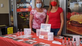 Two women in red tshirts stand in front of a red table covered with human trafficking information.