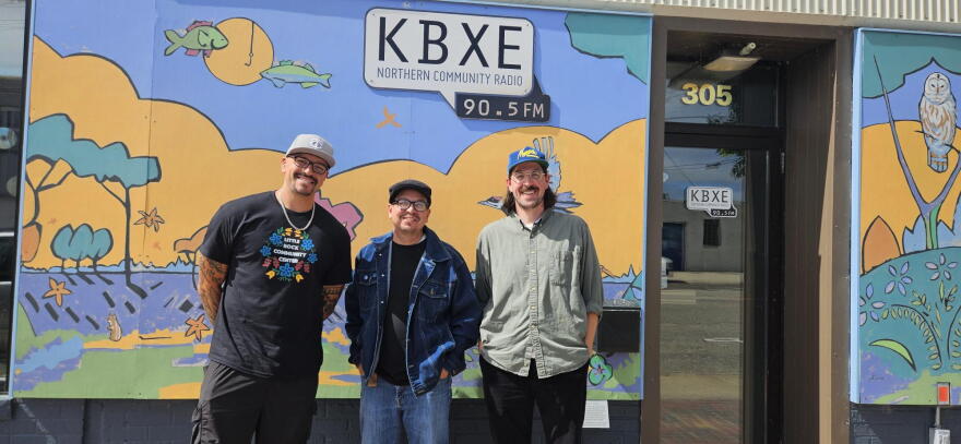 Three men standing in front of a building with a mural that says "KBXE Northern Community Radio 90.5 FM."