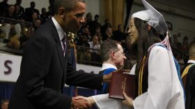 The photo is a close-up of a man in a black suit jacket and white undershirt shakes the hand of a woman in a white gown and matching white cap. They're standing on the stage in a large arena, with high up bleachers seen behind them.  