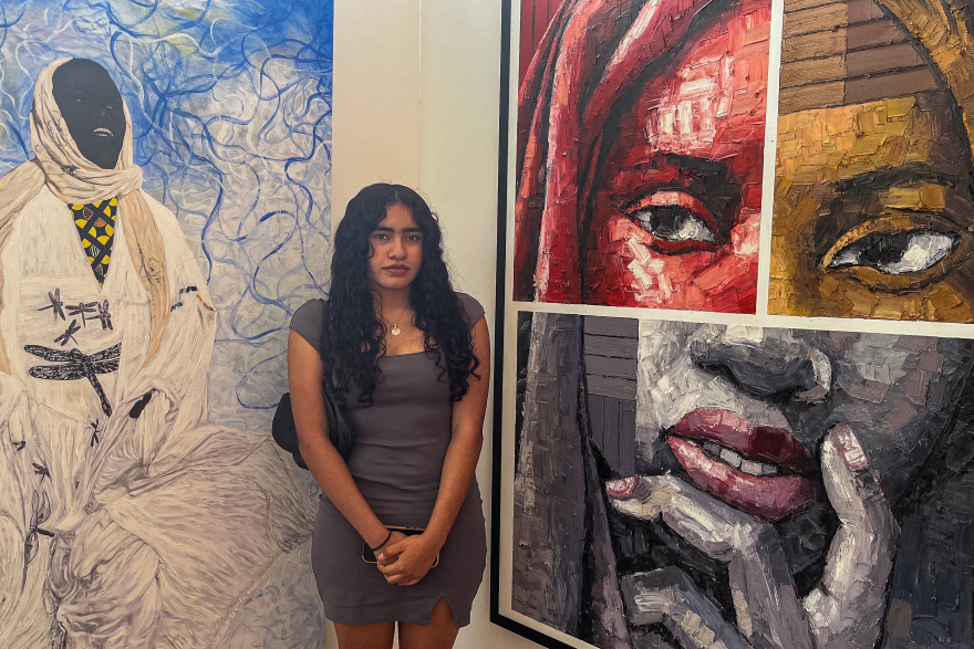 A young woman in a dress stands in front of paintings on the wall.