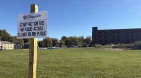 in the foreground, a white sign on a wooden post. in the background, an open field and brown brick building