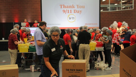 A woman wearing a hair net reaches for a tape roll with one hand as she places the other on a medium-sized cardboard box. Behind her, dozens of volunteers crowd tables. They pour rice, beans, and spices through funnels into bags. A screen on the back wall reads "Thank you 9/11 Day"