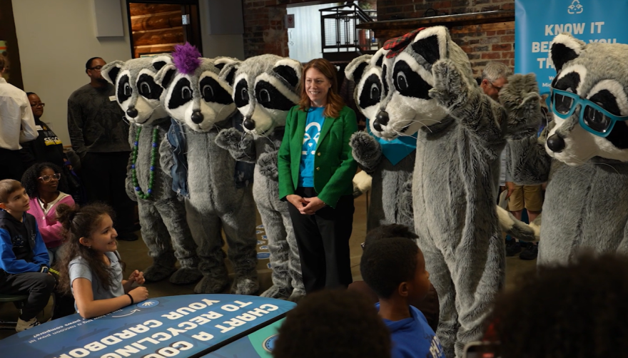 Tracy Kecskemeti smiling in a green blazer standing in the center of a group of six raccoon mascots. 

In front of them is a group of children seated, looking toward the mascots. A blue table sign in front of them reads, "Chart a course to your recycling," while a banner in the background says, "Know it before you throw it," at an indoor community event focused on environmental awareness.