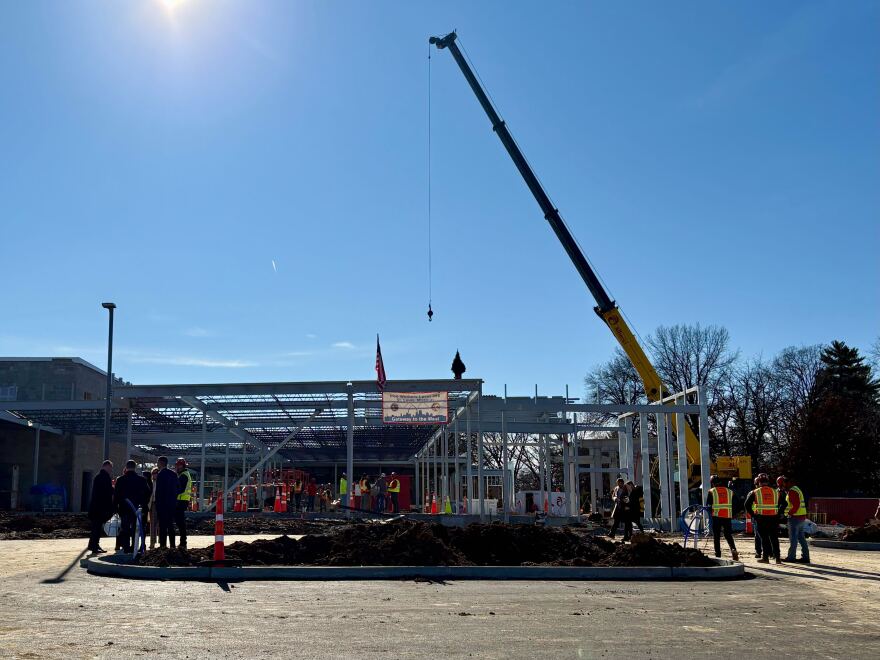 A wide shot shows the final beam in place at the under-construction mental health facility in Webster Groves. The horizontal beam is at the roof-level of the building, and workers have attached a small tree and flag to the the top. 