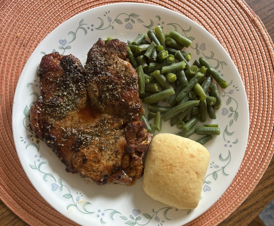 A plate with a pork chop, dinner roll and green beans is sitting on an orange placemat.