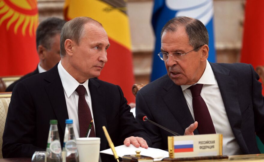 Putin and Lavrov speak while sitting next to each other at a table, with colorful flags in the background.