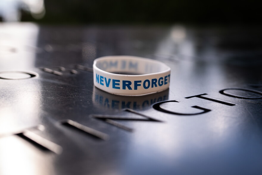 A white wristband with blue text reading “NEVER FORGET” rests on a reflective surface engraved with names, likely at a memorial site.