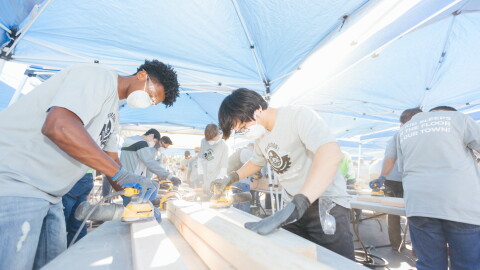 Volunteers wearing masks and safety glasses use power sanders on lumber beneath outdoor canopies.