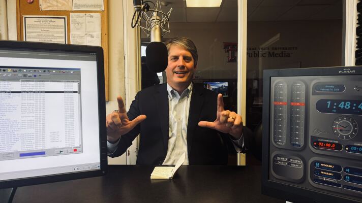 A photo of Tom Sell in the KTTZ studio. He is smiling and doing the Texas Tech 'guns up' hand sign.