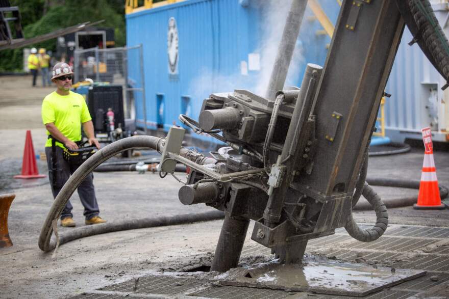 A man in a bright green shirt and hard hat operates a large machine that's drilling into the ground.