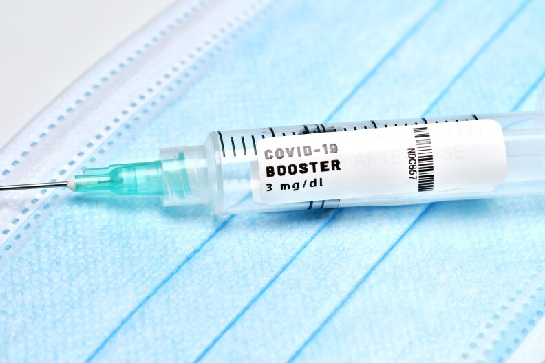 A syringe with a white label that says COVID-19 BOOSTER lays on a flat blue face mask.